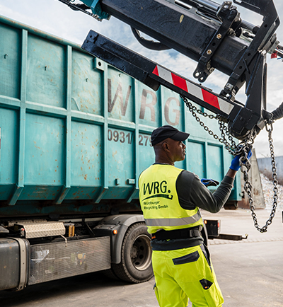 WRG – Containerverladung Mitarbeiter der WRG befestigt Container an einem LKW-Hubarm auf dem Recyclinghof.